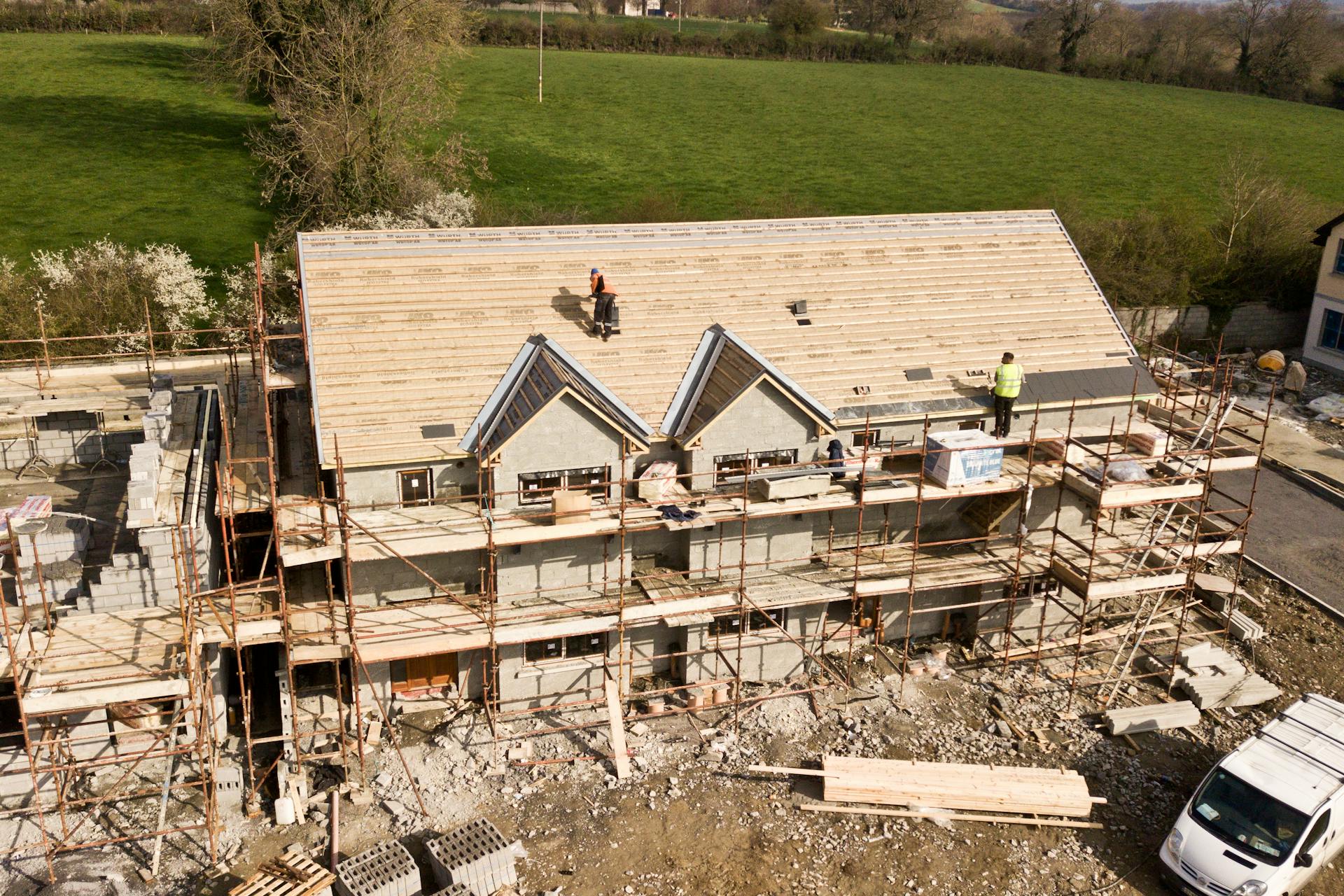 Construction workers building a large house with scaffolding and green fields in the background.