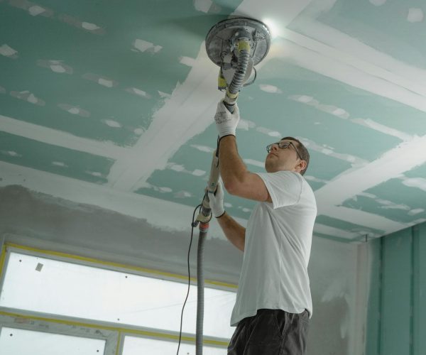Man using sanding machine on ceiling for smooth finish.
